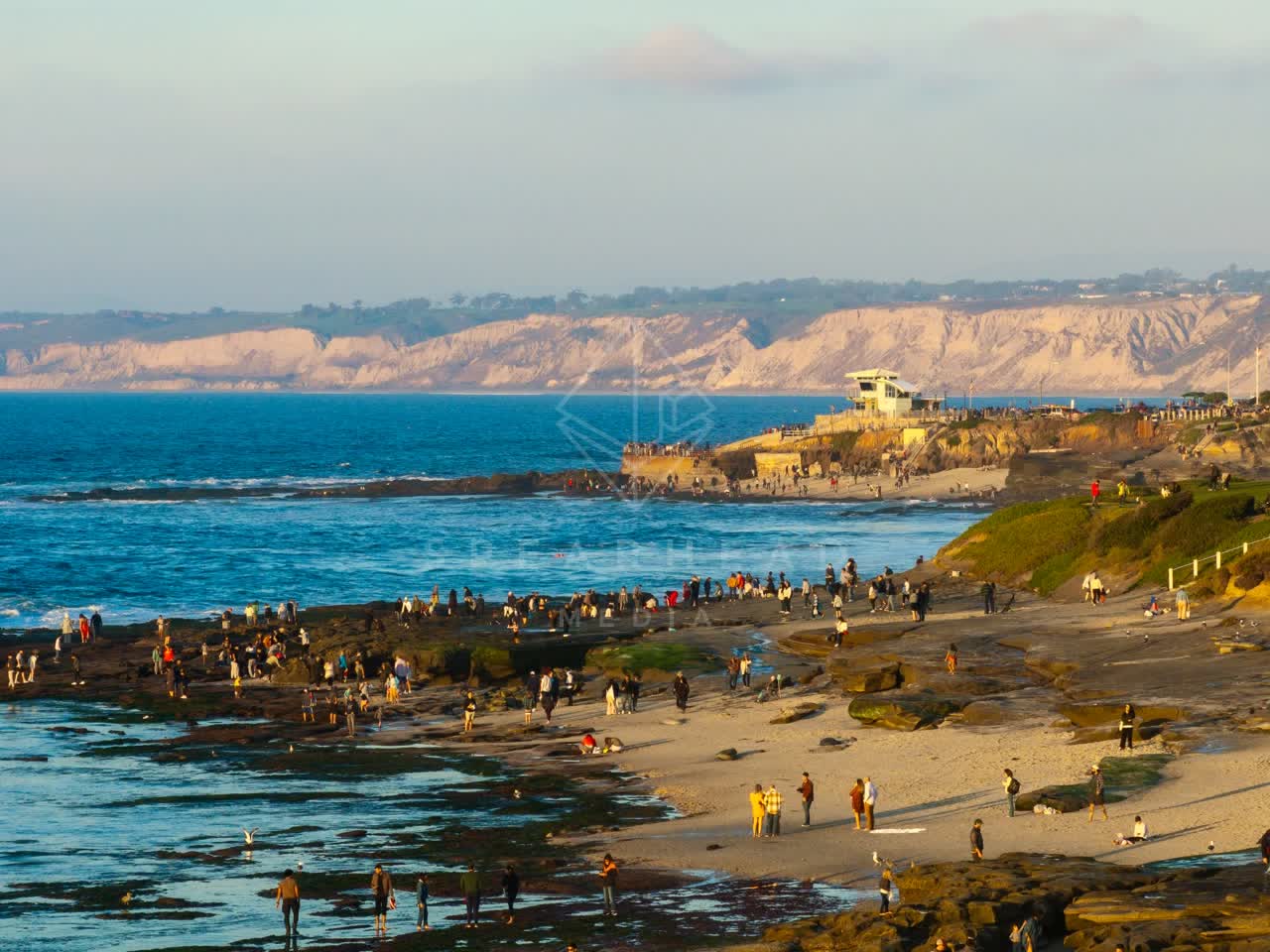 Low aerial view of the beach, the ocean and the cliffs at La Jolla Cove, with a crowd gathered.