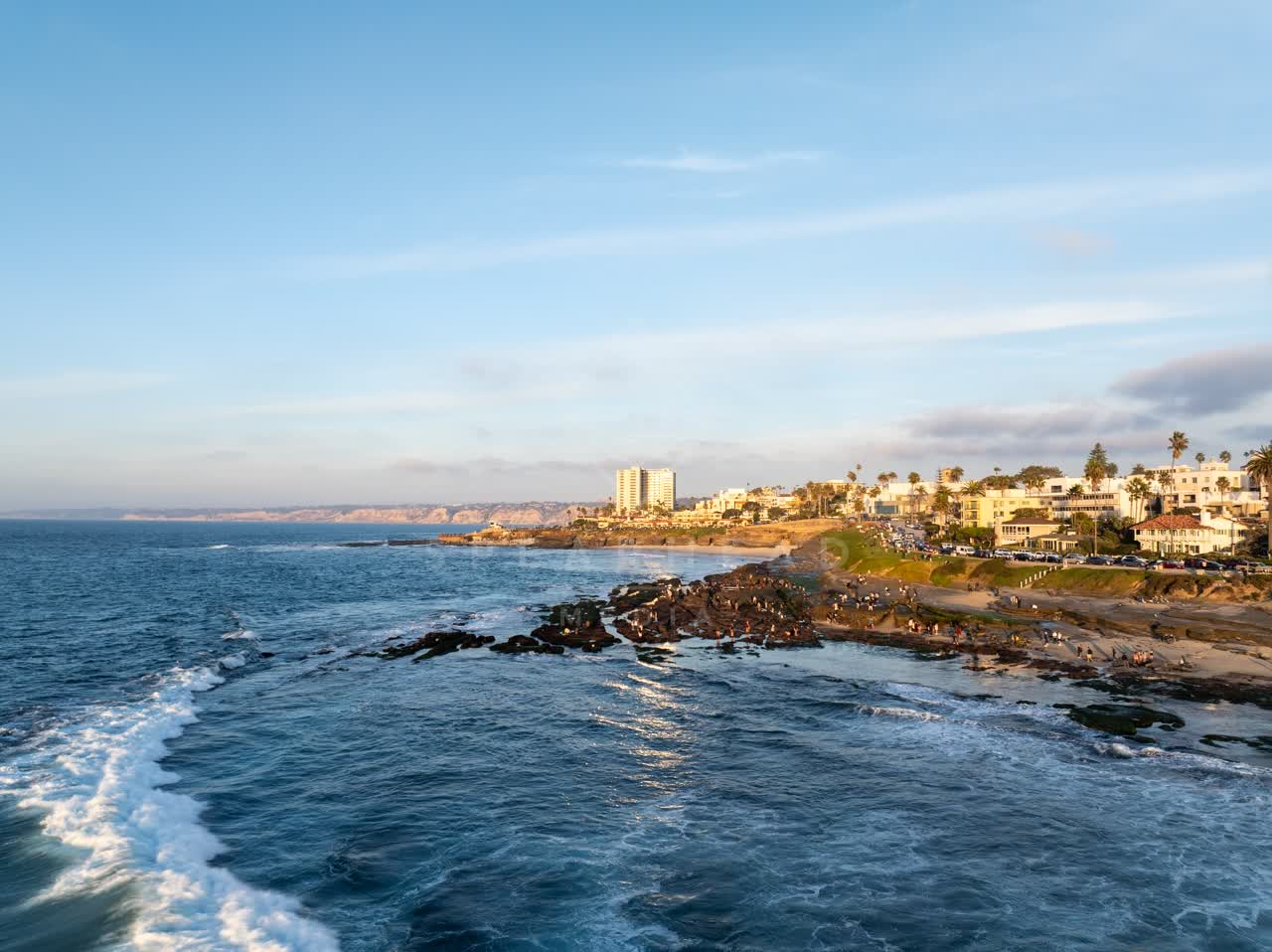 Aerial view of the cliffs, the surf and palm trees at La Jolla Cove, with a crowd gathered.