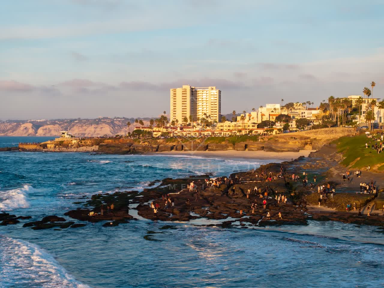 Low aerial view of the beach, the surf and palm trees at La Jolla Cove, with a crowd gathered.