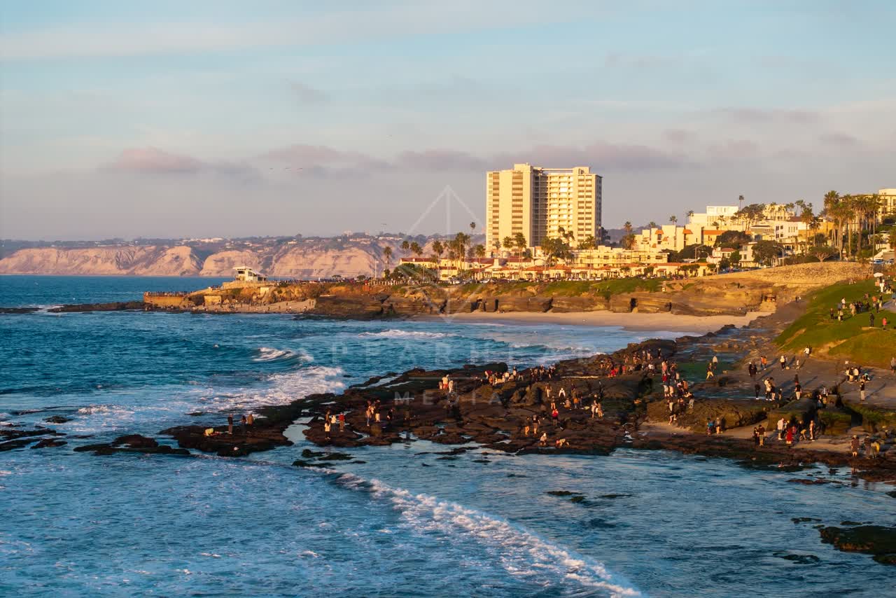 Low aerial view of the cliffs, the surf and the ocean at La Jolla Cove, with a crowd gathered.