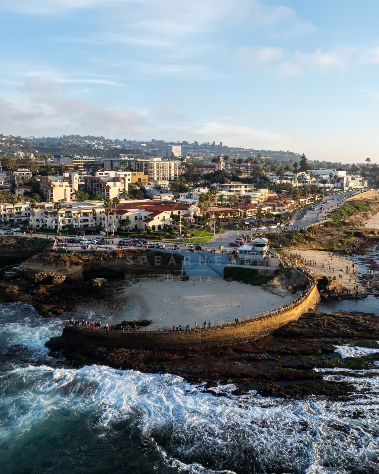 Aerial view of the cliffs, the surf and palm trees at La Jolla Cove, with a crowd gathered.