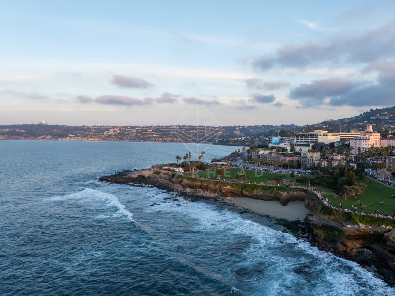 Aerial view of the cliffs, the surf and palm trees at La Jolla Cove, with a crowd gathered.