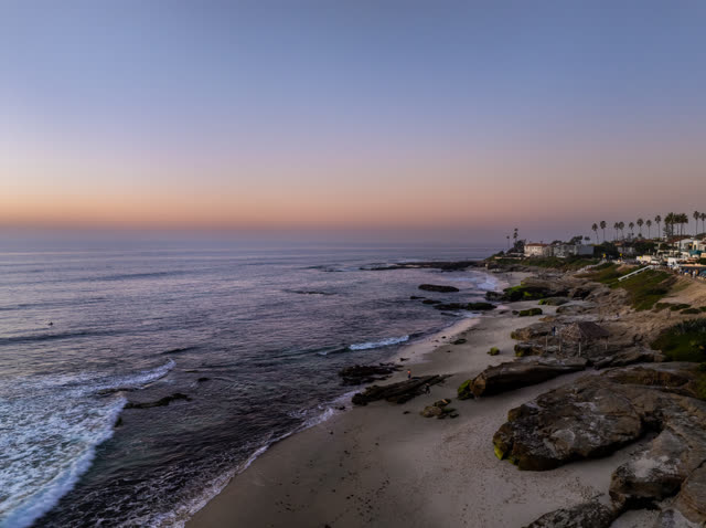 Low aerial view of the cliffs, the surf and palm trees at Windansea, with people walking.