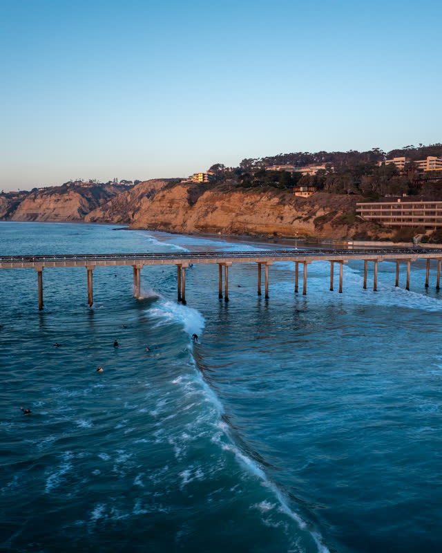 Low aerial view of the beach, the surf and the pier at La Jolla Shores, with surfers out.