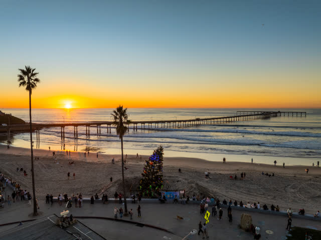 Low aerial view of the beach, the surf and the pier in Ocean Beach, with a crowd gathered.