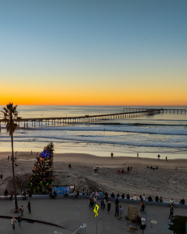 Low aerial view of the beach, the surf and the pier in Ocean Beach, with a crowd gathered.
