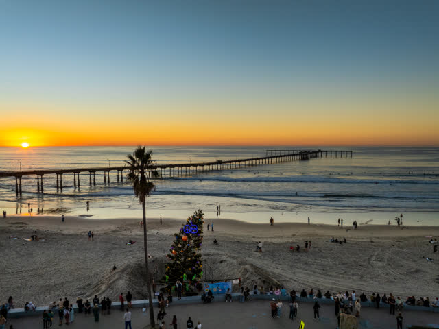 Low aerial view of the beach, the surf and the pier in Ocean Beach, with a crowd gathered.