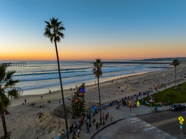 Low aerial view of the beach, the surf and the pier in Ocean Beach, with a crowd gathered.