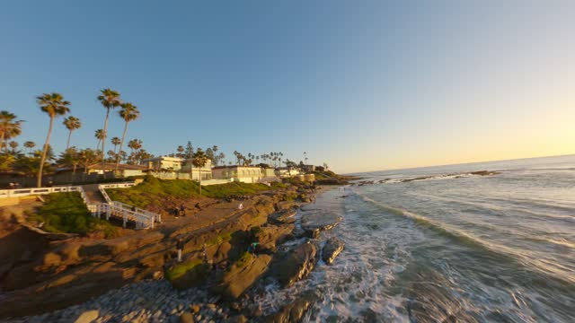 Low aerial view of the cliffs, the surf and palm trees at Windansea, with people walking.