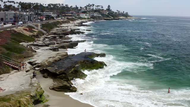 Low aerial view of the cliffs, the surf and palm trees at Windansea, with people walking.