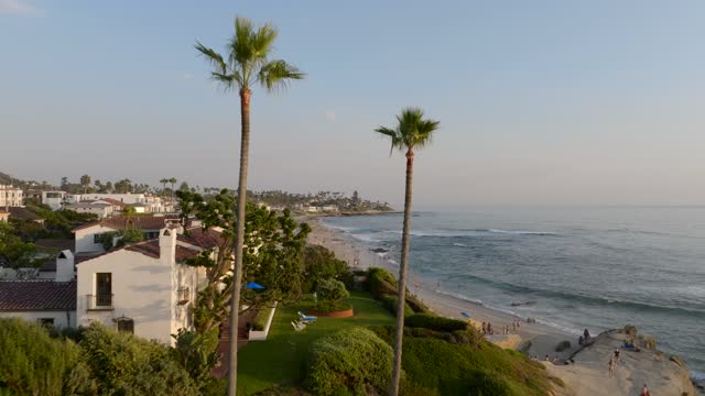 Low aerial view of a residential street, palm trees and the ocean at Beach Barber Tract, with people walking.