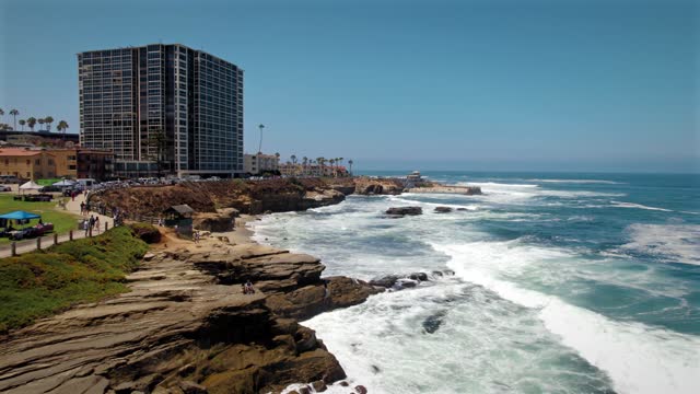 Low aerial view of the cliffs, the surf and palm trees at La Jolla Cove, with people walking.