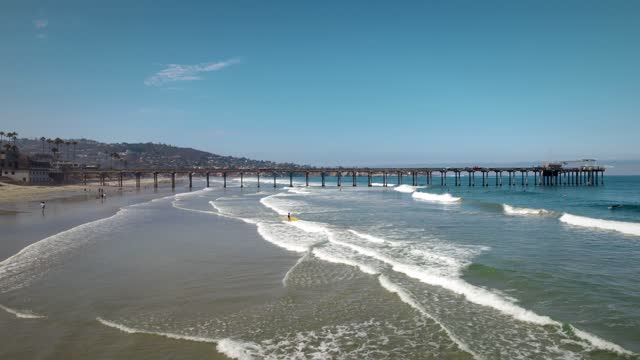 Low aerial view of the beach, the surf and the pier at La Jolla Shores, with surfers out.