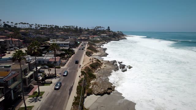 Low aerial view of a residential street, the surf and palm trees at Windansea, with people walking.