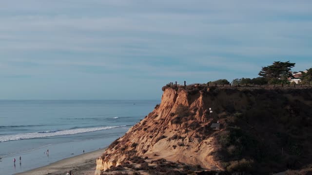 Low aerial view of the cliffs, the surf and the ocean at Del Mar Dog Beach, with people walking.