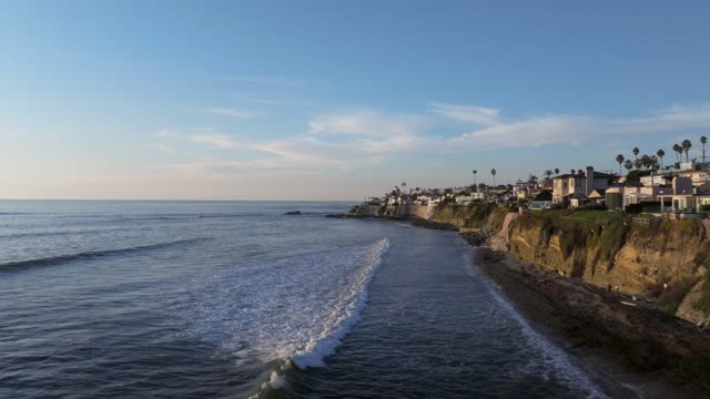 Low aerial view of the cliffs, the surf and palm trees at Bird Rock, with surfers out.