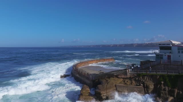 Low aerial view of the cliffs, the surf and the ocean at La Jolla Village, with people walking.