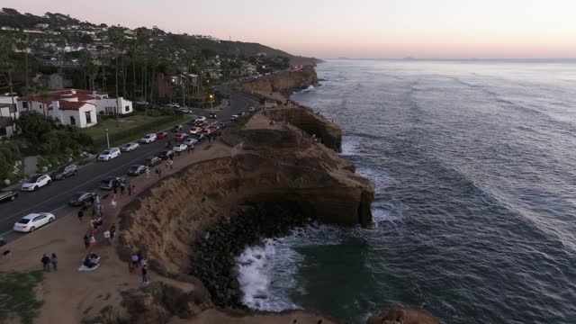 Low aerial view of the cliffs, the surf and palm trees at Sunset Cliffs, with a crowd gathered.