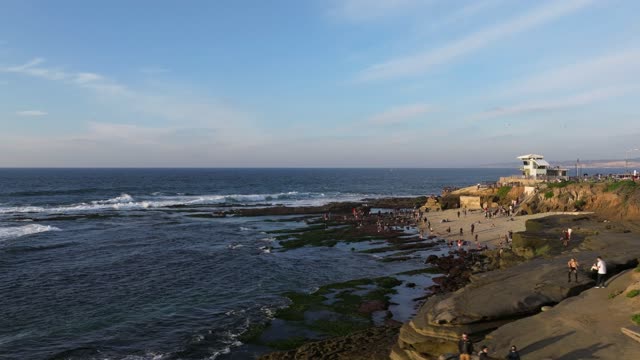 Low aerial view of the cliffs, the surf and the ocean at La Jolla Cove, with a crowd gathered.