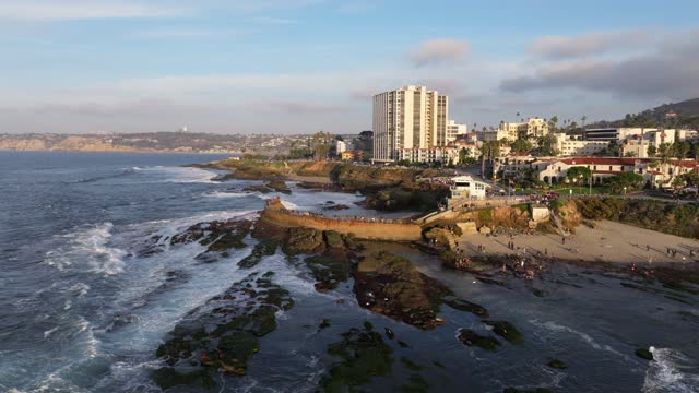 Low aerial view of the cliffs, the surf and palm trees at La Jolla Cove, with people walking.