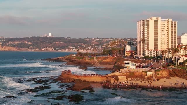 Low aerial view of the cliffs, the surf and palm trees at La Jolla Cove, with a crowd gathered.