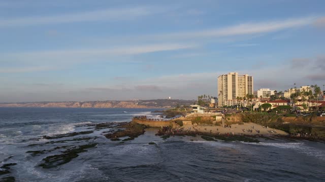 Low aerial view of the cliffs, the surf and wildlife at La Jolla Cove, with a crowd gathered.