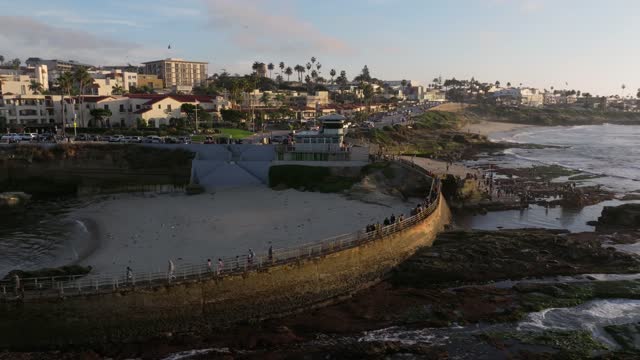Low aerial view of the cliffs, palm trees and the ocean at La Jolla Cove, with people walking.