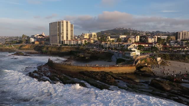 Low aerial view of the cliffs, the surf and the ocean at La Jolla Cove, with a crowd gathered.
