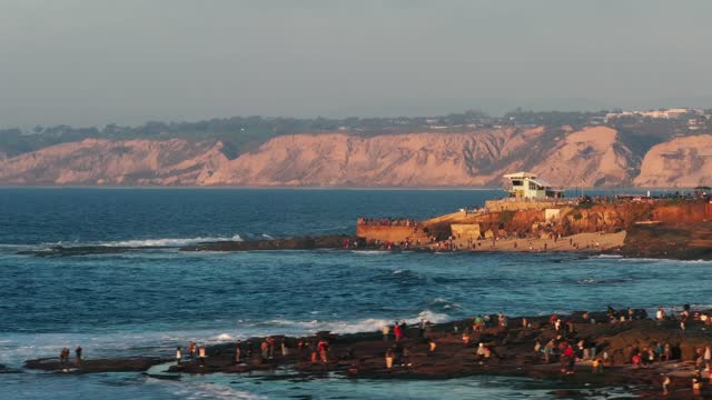 Low aerial view of the cliffs, the surf and the ocean at La Jolla Cove, with a crowd gathered.