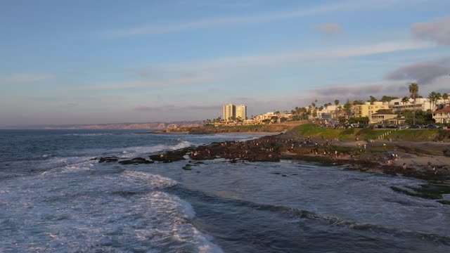 Low aerial view of the beach, the surf and palm trees at La Jolla Cove, with a crowd gathered.