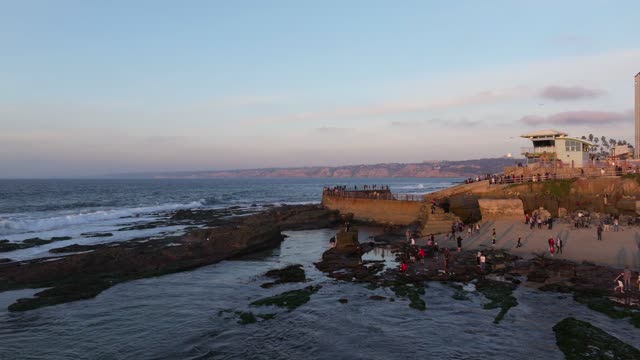 Low aerial view of the cliffs, the surf and the ocean at La Jolla Cove, with a crowd gathered.