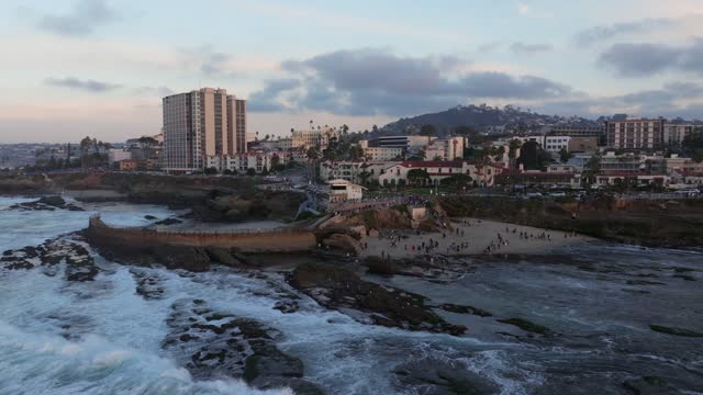 Low aerial view of the cliffs, the surf and palm trees at La Jolla Cove, with a crowd gathered.