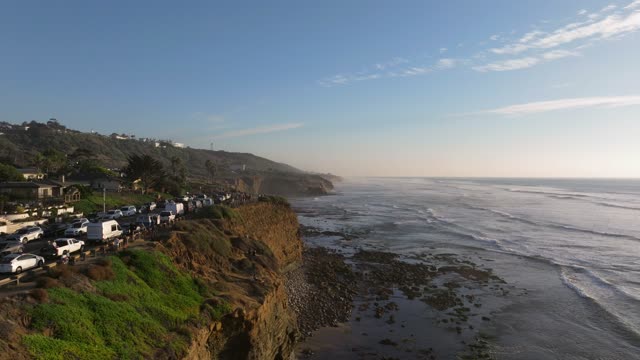 Low aerial view of the cliffs, the ocean and homes at Sunset Cliffs, with a crowd gathered.