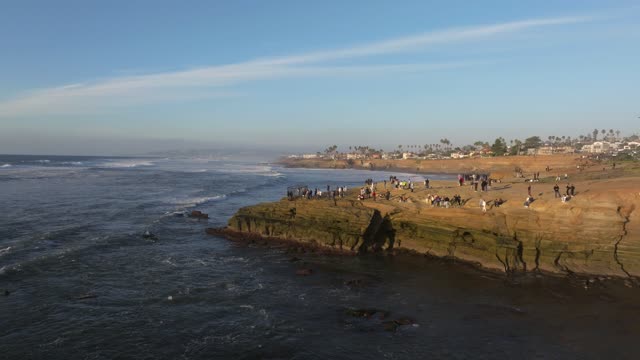 Low aerial view of the cliffs, the surf and palm trees at Sunset Cliffs, with a crowd gathered.