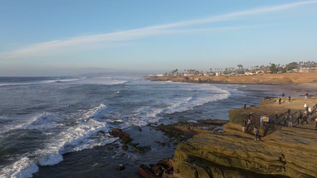 Low aerial view of the cliffs, the surf and palm trees at Sunset Cliffs, with a crowd gathered.