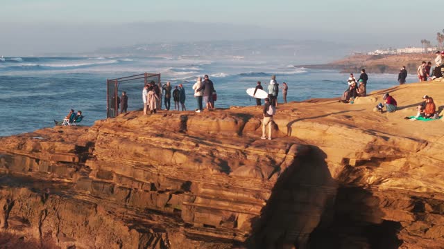 Low aerial view of the cliffs, the surf and the ocean at Sunset Cliffs, with a crowd gathered.