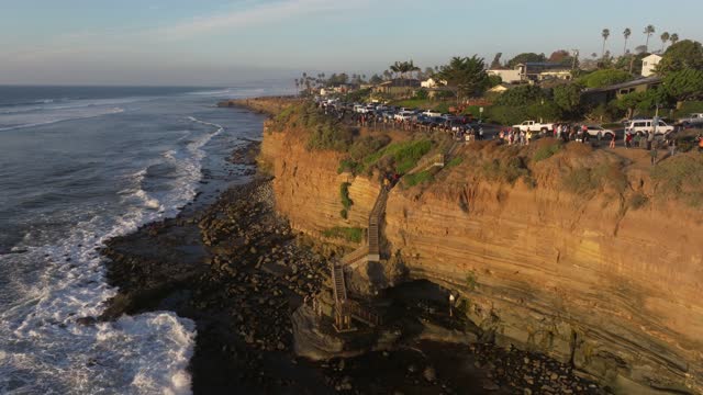 Low aerial view of the cliffs, the surf and palm trees at Sunset Cliffs, with a crowd gathered.