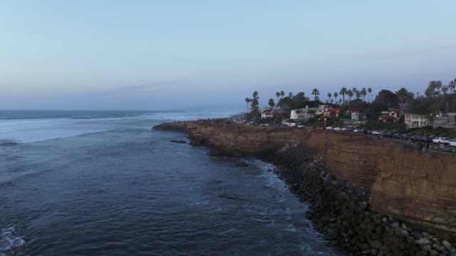 Low aerial view of the cliffs, the surf and palm trees at Sunset Cliffs, with a crowd gathered.