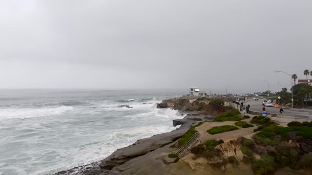 Low aerial view of the cliffs, the surf and the ocean at La Jolla Cove, with people walking.