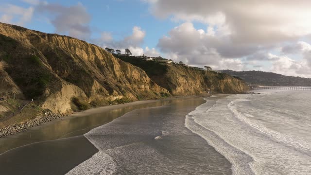 Low aerial view of the cliffs, the surf and the pier at Black’s Beach, with people walking.