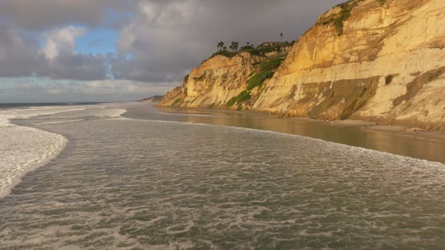 Low aerial view of the cliffs, the surf and palm trees at Black’s Beach, with people walking.