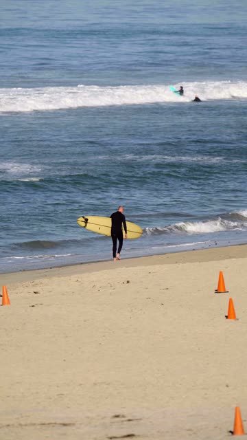 Surfer walking along the shoreline with a yellow surfboard as two surfers float beyond the waves at Moonlight Beach in Encinitas | Vertical Video