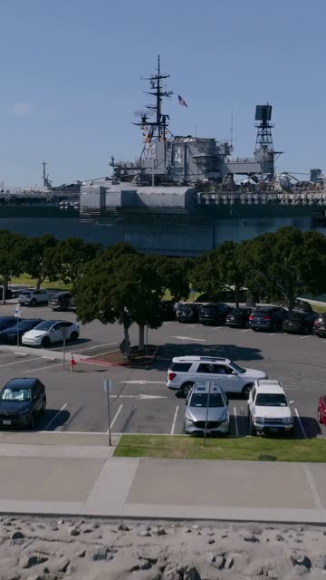 Aerial footage of the USS Midway Museum ship docked on the Embarcadero waterfront with parked cars and visitors in Downtown San Diego | Vertical Drone Video