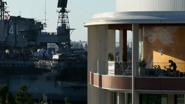 Aerial footage of the modern RADD building balcony with the USS Midway aircraft carrier docked at the Embarcadero in Downtown San Diego | Drone Video