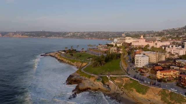 Aerial footage of La Jolla Cove, Scripps Park and La Valencia Hotel overlooking waves, cliffs and palm trees at golden hour | Drone Video