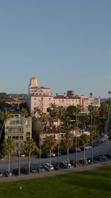 Aerial footage of the pink La Valencia Hotel, palm trees and Ellen Browning Scripps Park with waves breaking along the cliffs near La Jolla Cove | Vertical Drone Video