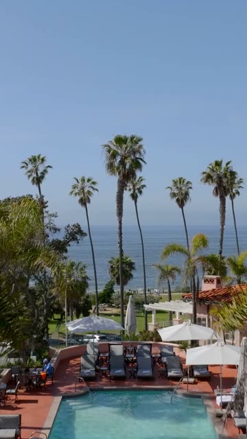Aerial footage of a hotel pool deck overlooking Scripps Park, palm trees and ocean waves at La Jolla Cove in La Jolla | Vertical Drone Video