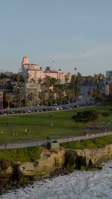 Aerial footage of the La Valencia Hotel and Ellen Browning Scripps Park with palm trees, cliffs and people walking at La Jolla Cove in La Jolla | Vertical Drone Video