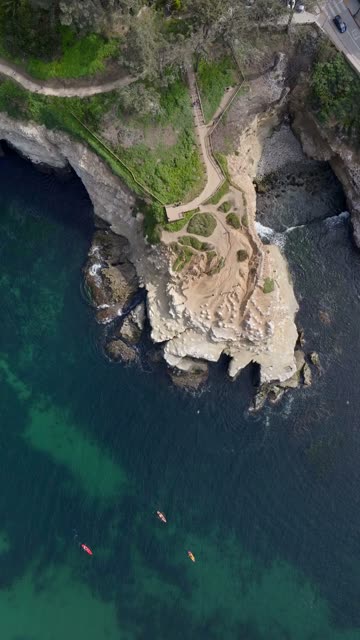 Aerial footage of kayakers paddling near a sandstone cliff peninsula and hikers on Coast Walk Trail above La Jolla Cove | Vertical Drone Video